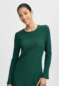 Robe longue à manches longues de couleur vert foncé fabriquée en tissu texturé avec une coupe légèrement ajustée et un col rond, présentant des motifs ondulés subtils à la surface.