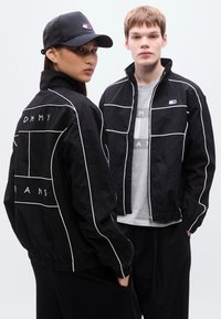 Μαύρα μπουφάν με λευκή περίγραμμα, με λογότυπο στο στήθος και στην πλάτη. Και ο ι δύο φορούν ταιριαστά μαύρα καπέλα· το φόντο είναι ουδέτερο.