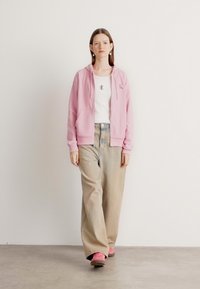 Felpa rosa con zip e tasche, indossata sopra una maglietta bianca. Pantaloni beige larghi e calzature rosa, in piedi contro uno sfondo chiaro.