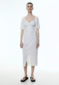 Weißes, knielanges Kleid mit quadratischem Ausschnitt, Puffärmeln und drapiertem Design; strukturierter Stoff; kombiniert mit schwarzen Sandalen.