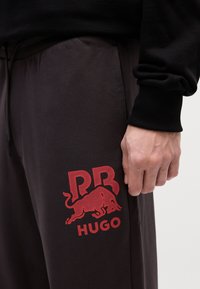 Tumši pelēki sporta bikses ar mīkstu faktūru, kas rotātas ar sarkano bulles grafisko attēlu un "RR HUGO" logo kreisajā augšstilbā.