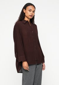 Camisa ligera de color marrón, tipo botón, con cuello, mangas largas y bolsillos frontales; presenta un ajuste relajado y una tela texturizada.