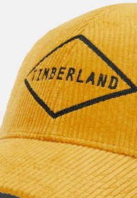 Gorra de béisbol de pana amarilla con un logo de Timberland bordado en negro en forma de diamante en la parte frontal. Se puede ver la textura rayada.