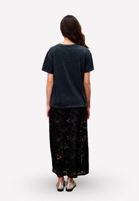 Frau mit langen Haaren, die ein verblasstes schwarzes T-Shirt, einen schwarzen Spitzmaxirock und silberne Ballerinas trägt, steht mit dem Rücken zur Kamera.