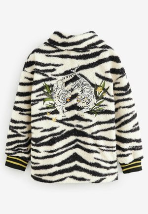 Veste en polaire blanche avec des rayures tigre noires. Présente des tigres brodés parmi des feuilles vertes et des étoiles. Manches noires avec des rayures jaunes.