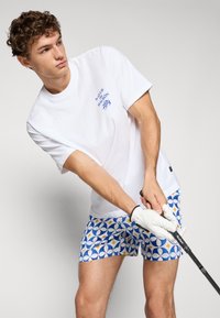 Hvid kortærmet t-shirt med blå tekst, parret med blå mønstrede shorts med gule accenter. Model holder en golfkølle.