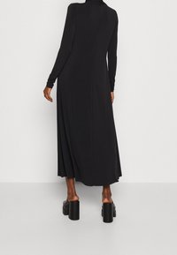 Musta pitkähihanen mekko, jossa on korkeakaulukko ja nilkkoihin ulottuva helma. Valmistettu sileästä kankaasta, rento istuvuus ja päällekkäin ommellut yksityiskohdat takana.