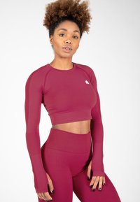 Bordeaux kleurige langemouwen sportieve crop top en hoog getailleerde leggings, gemaakt van rekbare stof met mesh-details op de mouwen en een klein logo op de top.