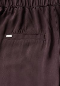 Mörklila tyg med en slät textur, featuring ett elastiskt midjeband och en liten metallmärkning som är sydd på fickan.