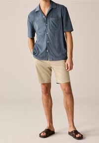 Lyhythihainen, sininen froteepaita rintataskulla, yhdistetty vaaleanruskeisiin shortseihin ja tummanruskeisiin liukusandaaleihin. Suoralahkeinen malli.