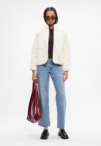 Giacca bianca in pile con due tasche frontali, abbinata a un top marrone scuro e jeans blu. Tiene una borsa a tracolla bordeaux lucida. Scarpe nere.