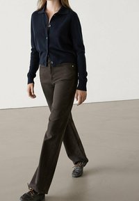 Granatowy kardigan z dzianiny z guzikami, w połączeniu z brązowymi spodniami typu slim-fit. Stylizacja uzupełniona czarnymi butami, przedstawiona na neutralnym tle.