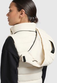 Verstärkter cremefarbener Weste mit hohem Kragen, ausgestattet mit einem schwarzen Reißverschluss und verstellbarem Kordelzug, kombiniert mit einem schwarzen Langarmtop.