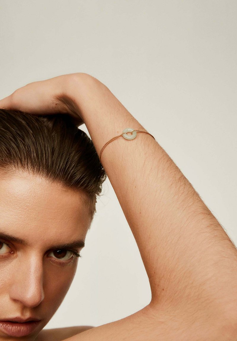 Pulsera de tono dorado con un centro de piedra circular y translúcida. El diseño es delicado con una fina cadena de metal y una textura suave.
