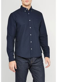 Donkerblauwe overhemd met lange mouwen en knopsluiting, voorzien van een kraag, een enkele borstzak, witte knopen en een rechte zoom. Draagt het met donkere spijkerbroeken.