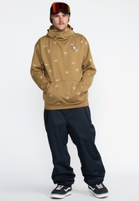 Sweat à capuche marron avec des motifs d'hirondelles, poche kangourou et un patch étoile ; associé à un pantalon de ski noir et des baskets.