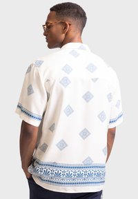 Chemise à manches courtes crème, ornée d'un motif géométrique bleu et d'une bande décorative le long de l'ourlet et des manches. Texture de tissu lisse.