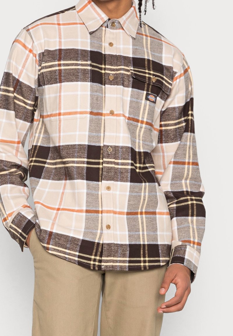 Homme portant une chemise à carreaux beige, marron et orange à boutons avec une poche poitrine et un pantalon beige, main dans la poche.