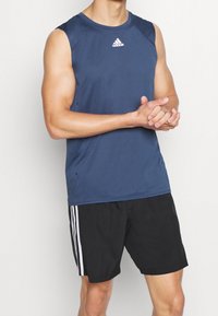 Tmavě modré sportovní tílko bez rukávů s tmavými panely na ramenou a logem na hrudi. Kombinováno s černými šortkami s bílými pruhy po stranách.