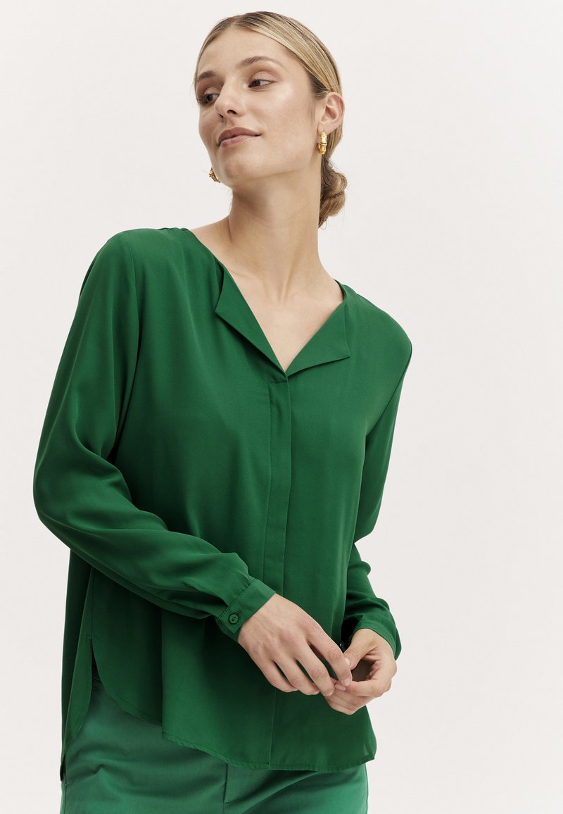 Green long-sleeve blouse with a v-neck, smooth texture, and buttoned cuffs. Features a slightly curved hem and a relaxed fit.