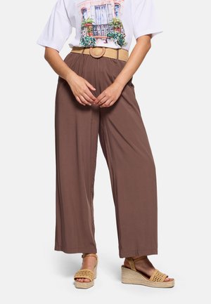 Mujer con pantalones marrones holgados con cinturón tejido, camiseta blanca con gráfico y sandalias cuña beige con los dedos abiertos.