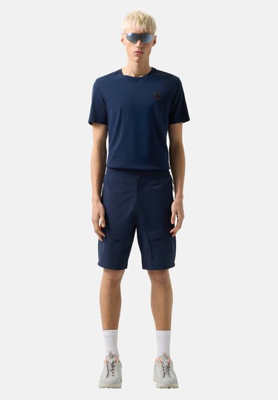 Personne blonde portant un t-shirt bleu foncé, un short cargo marine, des chaussettes blanches, des baskets grises et des lunettes de soleil enveloppantes réfléchissantes, debout face à l'avant.