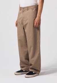 Beige wijde broek gemaakt van duurzaam materiaal, met een frontale knoopsluiting, zijzakken en rechte naden. G gedragen met zwarte sneakers.