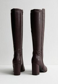 Botas altas hasta la rodilla de cuero marrón con tacón grueso, cremallera lateral, textura lisa y detalles mínimos de costuras.