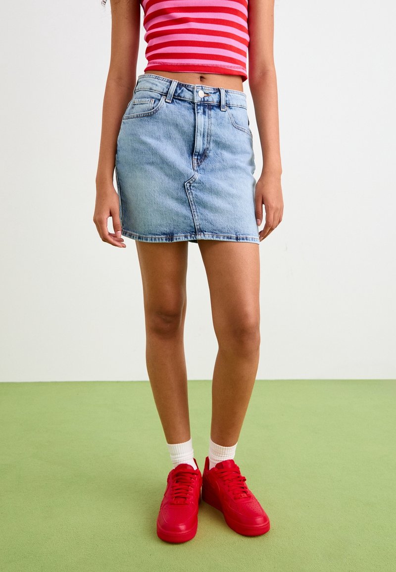 Personne portant un crop top rayé rouge et rose, une jupe en jean bleu clair, des chaussettes blanches et des baskets rouge vif, debout sur un sol vert.