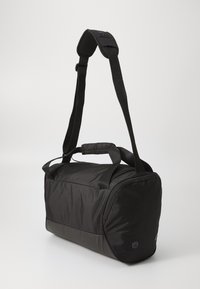 Svart stoff duffelbag med justerbar reim, doble topphåndtak, flat bunn og sideaksenter. Har en jevn tekstur og minimalistisk design.