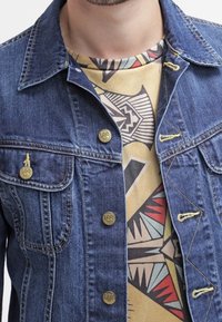 Denimjacka i mörkblått med gyllene knappar och gula sömmar, lager på lager över en mönstrad beige t-shirt med geometriska former.