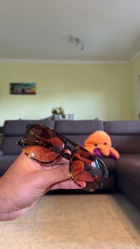 Γυαλιά ηλίου από ταρταρούγα σε ορθογώνιο σχήμα, με σκούρους καφέ φακούς και γυαλιστερούς σκελετούς. Κρατιόνται στο χέρι μπροστά από ουδέτερο φόντο.