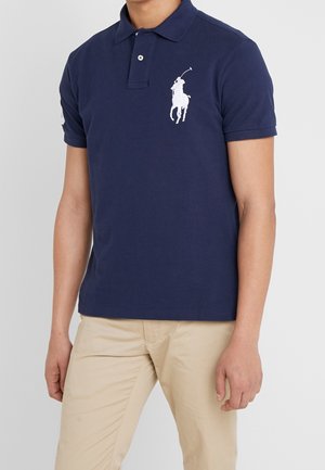 Man draagt een marineblauw poloshirt met wit logo van paard en ruiter, gecombineerd met beige broek, staand tegen een effen lichte achtergrond.