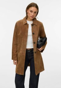 Pruun seemisnahast mantel kraega, millel viis eesmist nuppu ja kaks küljetaskut, kombineeritud valge topi ja tumedate teksapükstega. Käes must kott.