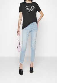 Camisa gráfica negra con un diseño alado en el frente, combinada con jeans de mezclilla claros y desgastados, y mules negros. Sosteniendo un bolso pequeño rosa.
