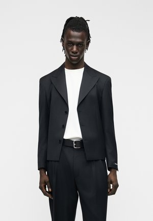 Άνδρας που φοράει μαύρο κοντό σακάκι πάνω από λευκό πουκάμισο, μαύρη ζώνη και μαύρο παντελόνι, στέκεται μπροστά σε απλό φωτεινό φόντο.