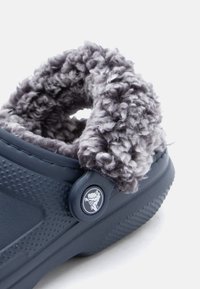 Tengerészkék, slip-on klumpa vastag szürke gyapjúbéléssel és texturált talppal, oldalán egy kerek emblémával.