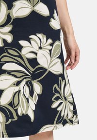 Blumenkleid aus navyblauem Stoff mit großen weißen und olivfarbenen Blüten. Das Design umfasst grüne Akzente und eine glatte Textur.