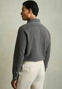 Cămașă gri din catifea cu un guler structurat și mâneci lungi, asociată cu pantaloni de culoare deschisă. Prezintă manșete cu nasturi și o textură netedă.