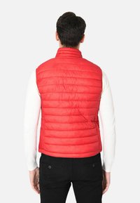 Gilet senza maniche trapuntato rosso con colletto alto, texture liscia e cuciture orizzontali, indossato sopra una maglietta a maniche lunghe bianca.