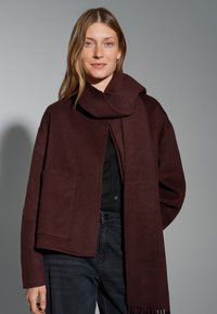 Burgundy wool blend scarf draped around the neck; oversized jacket with open front and two front pockets; worn over a black shirt.