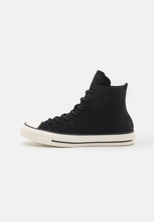 Baskets montantes noires avec semelle en caoutchouc blanche et bout renforcé, lacets noirs et design minimaliste sur fond gris clair.