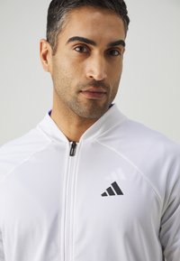 Witte sportieve zip-up jacket met een zwart Adidas-logo op de linkerborst, een gladde textuur, hoge kraag en een ritssluiting aan de voorkant.