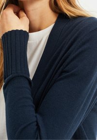 Marinblå cardigan med ribbstickade muddar och krage, med en mjuk textur. Buren över en vit skjorta, som visar en lager-på-lager-look.