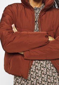 Doudoune couleur rouille avec un col montant, une texture matelassée et une fermeture éclair dorée, associée à une robe à motifs floraux en dessous.