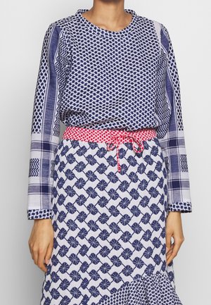 Femme portant un haut à manches longues bleu marine et blanc à motifs, avec une ceinture tressée rouge et blanche et une jupe à motifs floraux bleu marine.