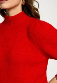 Comodo maglione rosso lavorato a maglia con un colletto alto e maniche leggermente a sbuffo. Tessuto morbido con un aspetto caldo e vibrante.