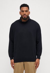 Schwarzer Strickrollkragenpullover mit langen Ärmeln, einem gerippten Kragen und einem lockeren Schnitt. Kombiniert mit beigen Hosen.