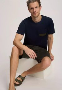 Granatowa koszulka z krótkim rękawem z zieloną kieszenią, zestawiona z ciemnymi szortami i brązowymi sandałami. Model siedzący na tle białego sześcianu.