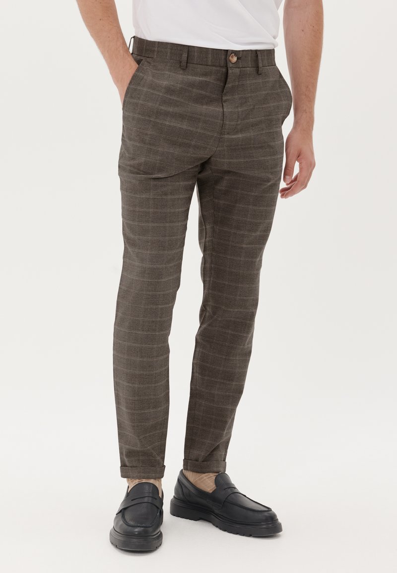 Braune Karohose aus strukturiertem Stoff, mit flacher Vorderseite, zwei Seitentaschen und einem Knopfverschluss. Getragen mit schwarzen Schuhen.